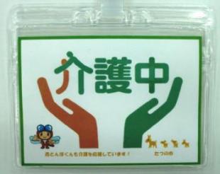 介護中という大きな文字と、その文字を包み込むようなオレンジ色の手と緑色の手のマークが載った名札の写真