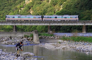 川にかけられた鉄道の橋の上を走る列車と、そのそばの川で釣りをする男性を横から写した作品