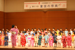 様々な色の浴衣を着てステージの上で合唱をする子どもたちと、子どもたちの向かい側に立って合唱の指揮を執る浴衣姿の女性を写した写真