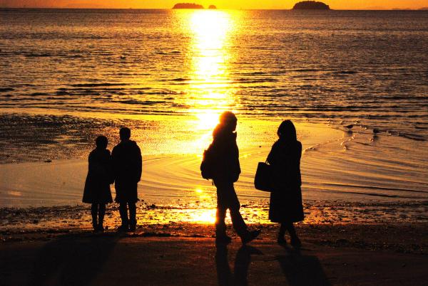 薄暗い中太陽の光で金色に染まった海と空と、水平線から砂浜の海岸へ続く太陽の光の道、その海や太陽を砂浜から眺める四人の人々を後方から写した写真