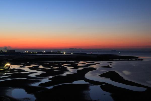 暗い色から赤っぽいオレンジ色に変わる夜明けの空と、潮が引いて干潟ができた遠浅の砂浜を写した作品