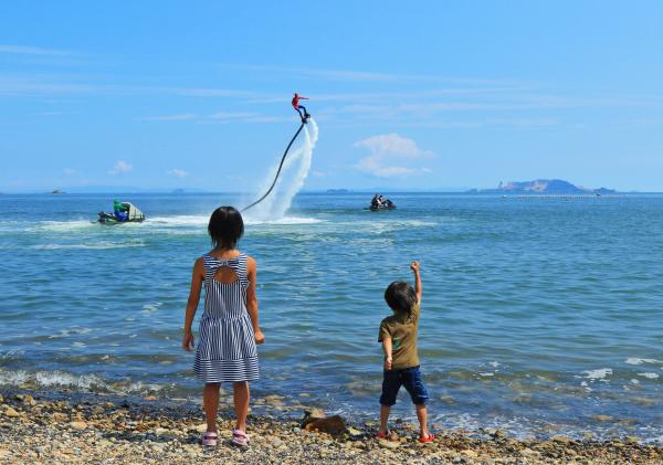 水上バイクから噴出される水圧を利用してかっこよく海の上を飛ぶ人と、その人を浜辺から夢中で見つめる女の子と小さな男の子を後ろから写した作品