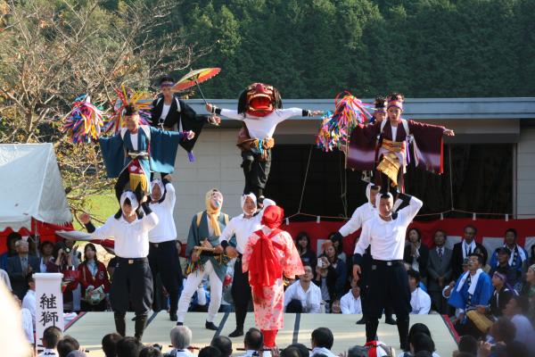 観客たちの前で、獅子頭をつけた人やカラフルな飾りを持った人が男性たちの肩の上に乗ってアクロバットな演技を披露している写真