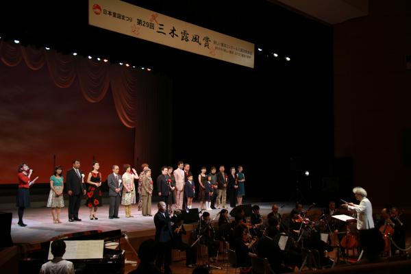 ステージのそばで様々な楽器を演奏する楽団の人々と指揮者と、胸にリボンのバラをつけてステージの上に横並びになった人々を斜め前から写した写真