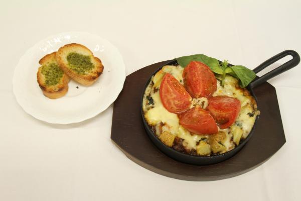 スキレットに入ったカレーにチーズが溶け真ん中に四等分したトマトの焼きカレーと白いお皿にバジルソースが塗られた2切れのバッケットが添えられた写真