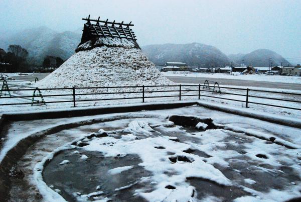 古代の竪穴住居の建物と、手前にある円形の遺跡の跡に雪が降り積もった様子を写した作品