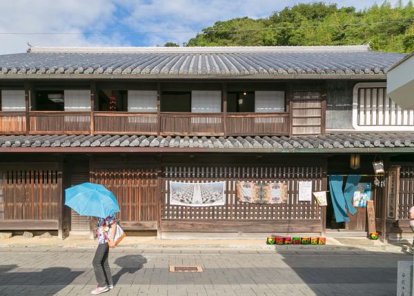 歴史を感じる瓦屋根の二階建ての建物と、その建物の前を水色の日傘をさして歩く人を写した作品