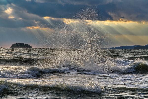 砕けて白いしぶきをあげる海の波と、雲のすき間からいくつもの光の筋がさす空を写した作品