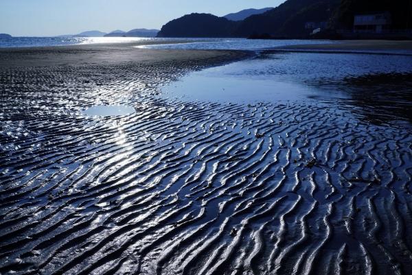 潮が引いて、さざ波のような跡がついた砂浜の上に残った海水や、海の表面がやや黒みを帯びた青色に染まっている様子を写した作品