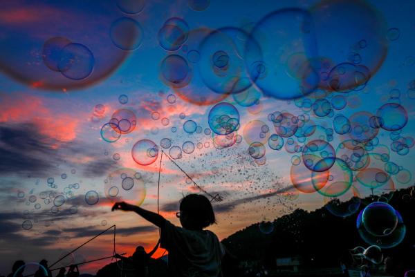 暗くなった青空や茜色の太陽の光を反射した雲が美しい夕暮れに、たくさんのシャボン玉を飛ばす子どもを斜め後方から写した作品