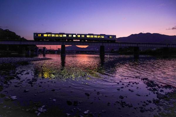 陽が沈みかけた山を背景に鉄橋を走っている電車を川岸から撮影した写真