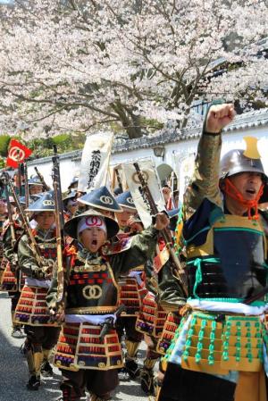 兜をかぶった鎧姿の男性と、そのあとに続く円錐形の帽子を被った鎧姿の子どもたちの行列を写した写真