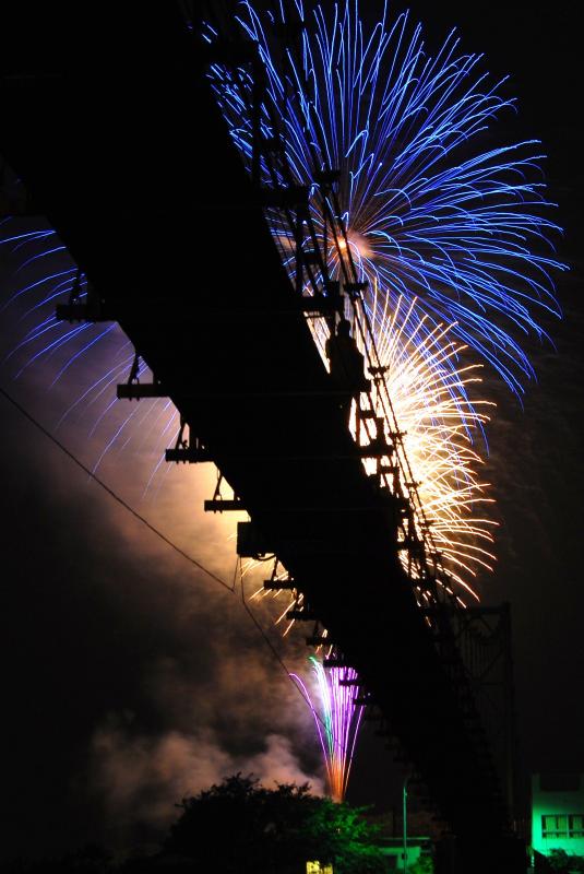 暗い夜に下から見上げた真っ黒で細長い吊橋の裏側と、その吊橋の向こうの夜空に打ちあがる青や金色の大きな花火を写した作品