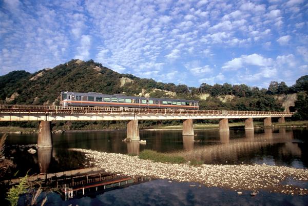 川にかけられた鉄道の橋の上を走る列車と、ひつじ雲のような雲がたくさん浮かんだ青空を斜め前から写した写真