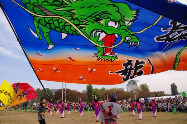 口を大きく開いた勇ましい龍と空を舞う赤とんぼの絵が描かれたとても大きな旗を振る男性と、その男性の奥で紫とピンクの衣装を着て何かの踊りを踊る人々を斜め後方から写した作品