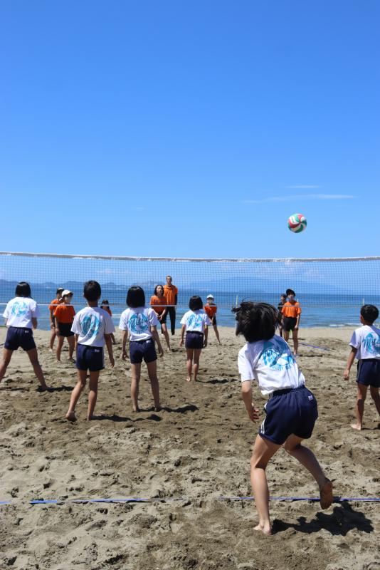 ネットを張った砂浜のコートでビーチバレーの対戦をする運動着姿の子どもたちを後方から写した写真