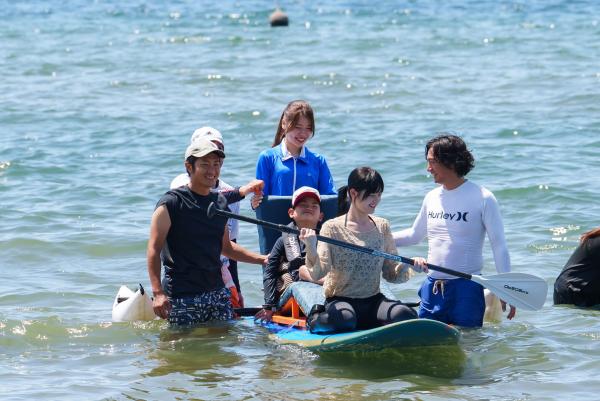4人のスタッフが見守る中、キャップを被った男の子と女性が海の上でSUP体験を行っている様子の写真