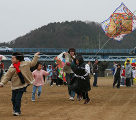 河川敷で大きな凧を楽しそうにあげる子どもたちと、子どもたちに付き添う大人たちを写した写真