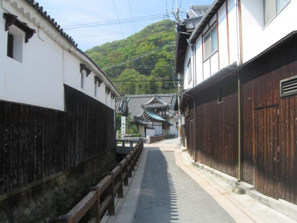 町中の小道沿いに建つ濃い茶色の板と白壁の外壁の建物や、その先に見える瓦屋根の立派な建物の写真