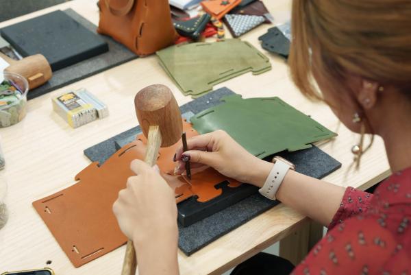 キャメルと緑色の革を重ねた上から、女性が道具を使用し穴をあけようとしている革細工ワークショップ体験を行っている様子の写真