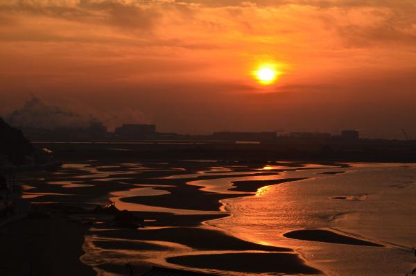 潮が引いて干潟になった遠浅の砂浜と、空に浮かぶ雲をオレンジ色に染める太陽の写真
