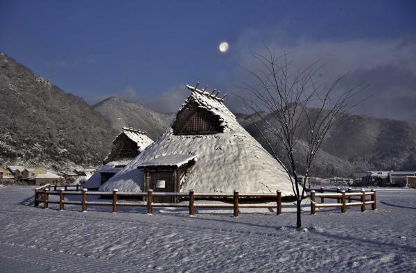 薄暗い空に浮かんだ白い月と、雪の積もった竪穴住居を写した作品