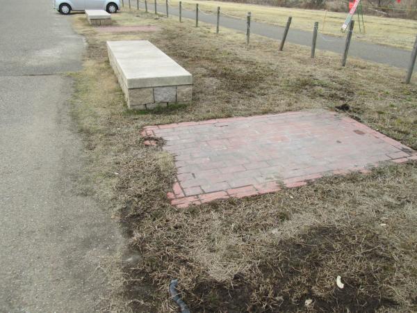 道路に面している石製の土台とレンガのエリアのある芝生のバーベキューサイトの写真
