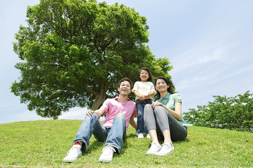 自然豊かな芝生が生えた丘で、地面に座った父親と母親の間に小さな女の子が笑顔で立っている親子の写真