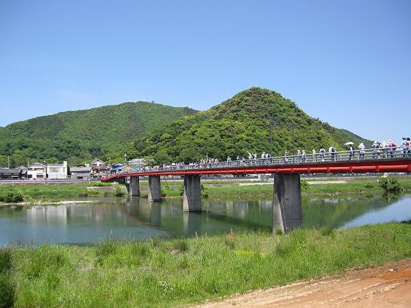 青空のもと、川にかけられた大きな橋を歩いて渡る大勢の人々を橋のそばの川岸から写した写真