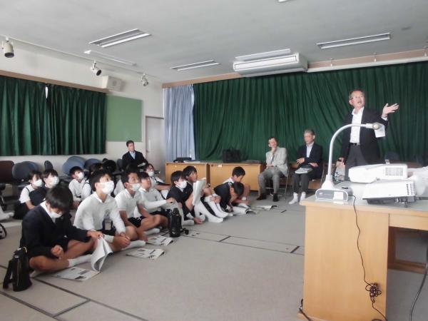 カーテンが閉められた室内で先生の説明を床に座り聞いている生徒たちの写真