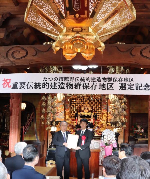 選定記念の幕が飾られた重要伝統的建造物の下で男性が2名で選定書を持ちながら記念撮影をしている写真