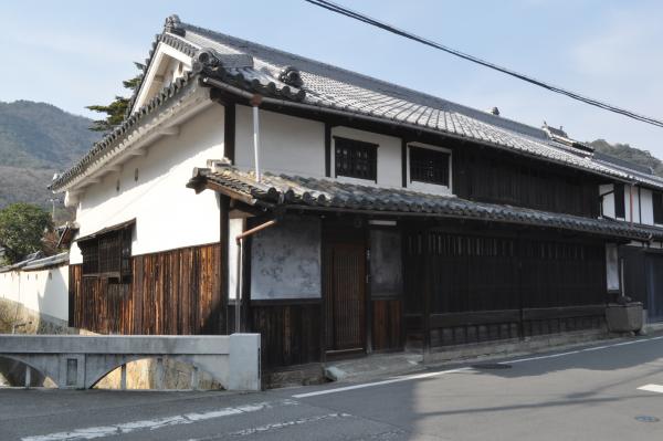 水路と道路が交差する場所に残る、窓に格子がはめられた白壁の古民家の建物外観の写真