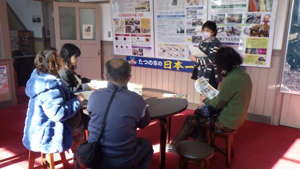 4名の参加者の人たちが丸いテーブルに座りパネルの説明をしている女性の話を聞いている写真