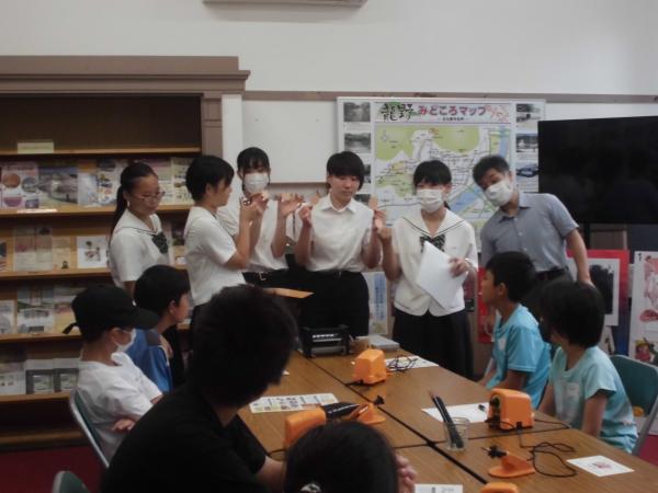 制服姿の高校生5人と男性が前に立ち説明をし長机を向かい合わせにした机に、はんだこてや説明の用紙が並べられ参加者が6人が座って話を聞いてる写真