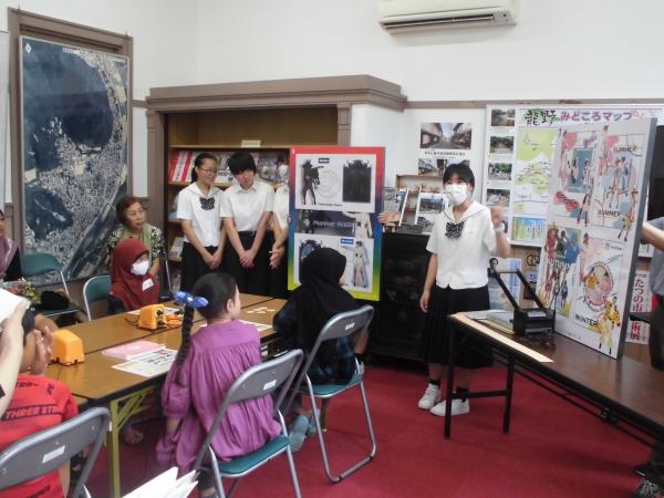 高校生がファッションショーの写真や説明文が載っているパネルを使い前に立ち話しているのを参加者が聞いている写真