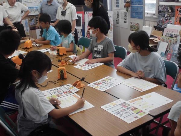 長机を向かい合わせにした机にはんだごてや資料が置かれ、用紙にデザインを描いている参加者の写真