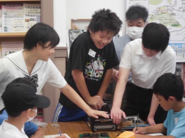 男子児童が型抜きの機械を笑顔で押し両側で高校生が支えている写真