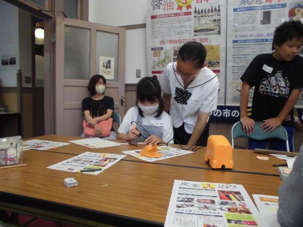 女子児童がはんだごてを持ち絵付けをしているのを後ろから高校生が見守っている写真