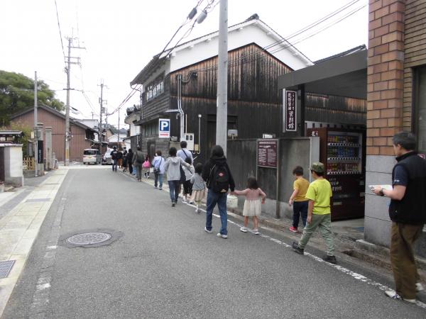 2階建ての和風の家が見える直線の道路を右側に縦一列に並んで歩く参加者の写真