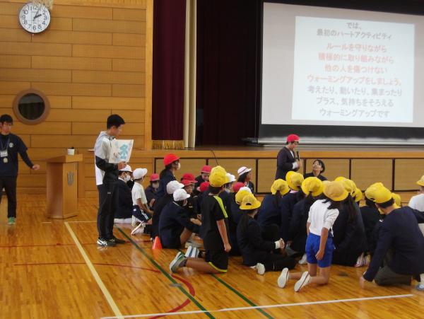 スクリーンに文章が映し出された体育館で、赤の帽子を被った児童が赤、白、黄、それぞれの帽子を被った児童たちの前でマイクを持ち話をしている写真