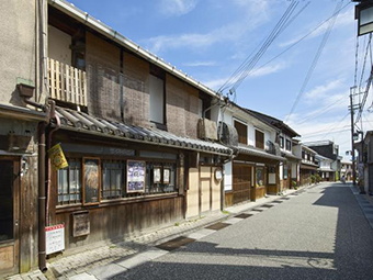 細い道路が奥まで真っすぐ伸びた左側に、窓に格子が設置され白壁で2階建ての町家造りの建物が建ち並んでいる町並みの写真
