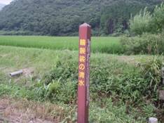 青々とした緑が生い茂っている背後に山や田んぼを背景に、黄色で「輪袈裟の清水」と書かれている赤色の四角い棒の看板のようなものを写した写真