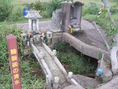青々とした緑の生えている土地に、石組の土台に花の供えられている二つの祠とその脇に黄色で「輪袈裟の清水」と書かれている赤色の四角い棒の看板のようなものを写した写真