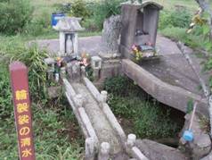 青々とした緑の生えている土地に、石組の土台に花の供えられている二つの祠とその脇に赤色の四角い棒の看板のようなものに黄色で「輪袈裟の清水」と書かれている写真