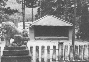 林に囲まれ松尾神社境内に設置された狛犬の奥に建つ、平屋建ての農村舞台の建物外観のモノクロ写真