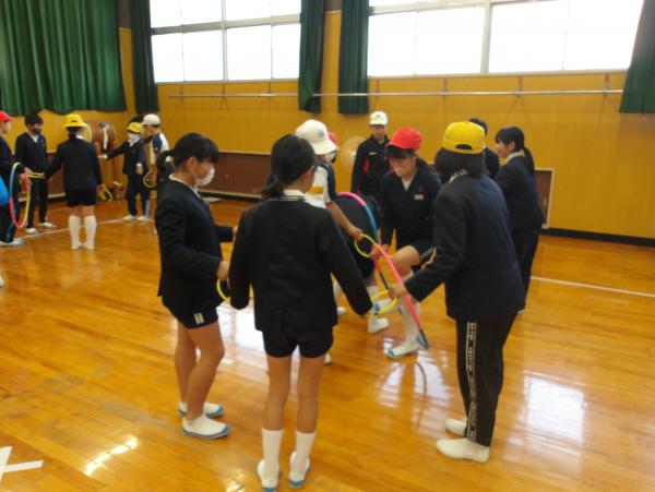 体育館でリングを手で握って、円形になった児童たちが大きなフラフープを使ってゲームをしている写真