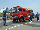 止められた消防車の前に立ち右手を上げている隊員と、消防車のホースを接続した部分の後ろで右手を上げている隊員の写真