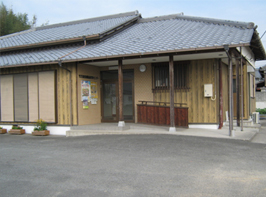 脇のほうに花の植えられたプランターの置かれた、木製の外観と灰色の瓦屋根の竹万農業構造改造センター