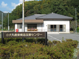 青空の下木々の生えた山を背景に映る、手前の植え込みに看板がある白い外観と灰色の瓦屋根の小犬丸農業構造改造センターの写真