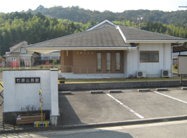 青々とした山を背景に映る、白色の塀に表札のある白と茶色の外観の竹原公民館の写真
